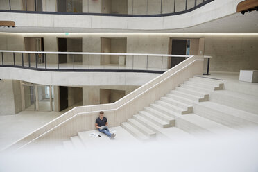 Young man sitting on stairs in office building using laptop - FMKF002989