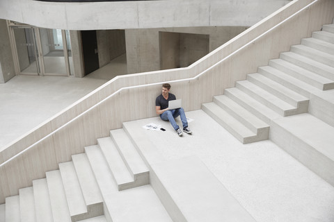 Junger Mann sitzt auf einer Treppe in einem Bürogebäude und benutzt einen Laptop, lizenzfreies Stockfoto