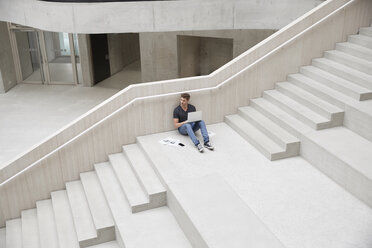 Junger Mann sitzt auf einer Treppe in einem Bürogebäude und benutzt einen Laptop - FMKF002990