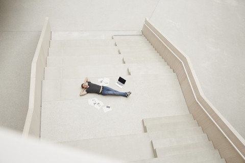 Young man lying on staircase with laptop and papers stock photo