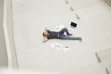 Young man lying on staircase with laptop and papers - FMKF002997