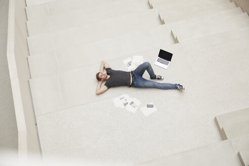 Junger Mann auf der Treppe liegend mit Laptop und Papieren - FMKF002997