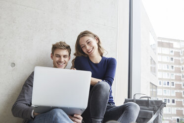 Smiling young couple sitting at concrete wall sharing laptop - FMKF003005