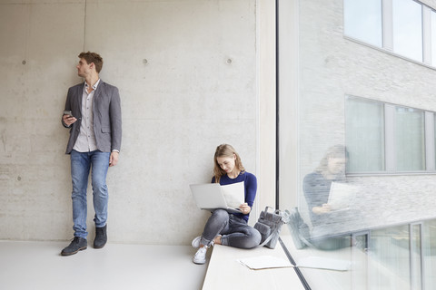 Junge Frau mit Papieren und Laptop und Mann mit Handy an Betonwand, lizenzfreies Stockfoto