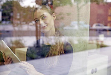 Junge Frau hält ein Tablet und schaut aus dem Fenster - FMKF003021