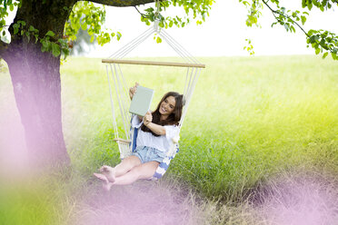Happy woman taking selfie with tablet in a hanging chair under a tree - MAEF011950