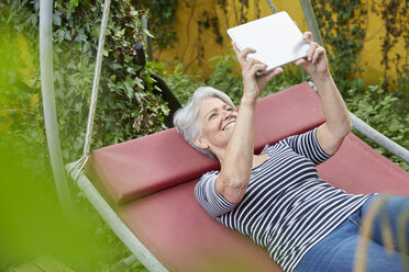 Frau entspannt sich auf einer Schaukel im Garten und benutzt ein digitales Tablet - FMKF003027