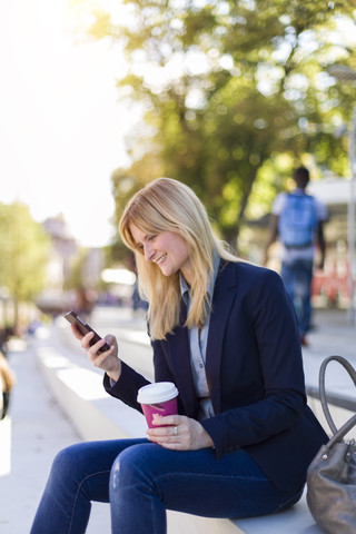 Lächelnde Geschäftsfrau mit Kaffee zum Mitnehmen, die auf ihr Smartphone schaut, lizenzfreies Stockfoto