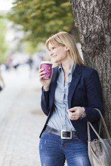 Smiling blond businesswoman with coffee to go leaning against tree trunk - NAF000036