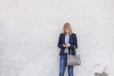 Lächelnde blonde Geschäftsfrau steht vor einer Wand und schaut auf ihr Smartphone - NAF000040