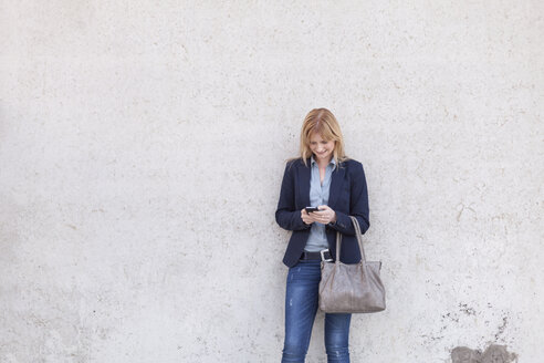 Smiling blond businesswoman standing in front of wall looking at smartphone - NAF000040