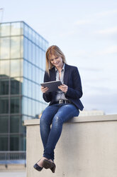 Lächelnde Geschäftsfrau sitzt an einer Wand und schaut auf ein Tablet - NAF000050
