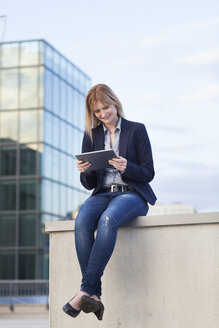 Smiling businesswoman sitting on a wall looking at tablet - NAF000050