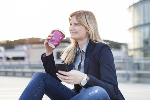 Lächelnde blonde Geschäftsfrau mit Smartphone und Kaffee zum Mitnehmen, lizenzfreies Stockfoto