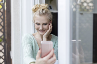 Blonde Frau sitzt an der Terrassentür und hält ein Handy in der Hand - SHKF000665