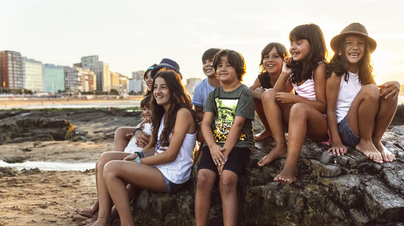 Eine Gruppe von Kindern sitzt bei Sonnenuntergang auf einem Felsen am Strand - MGOF002311