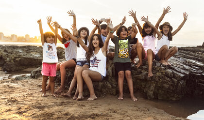 Eine Gruppe glücklicher Kinder sitzt bei Sonnenuntergang auf einem Felsen am Strand - MGOF002312
