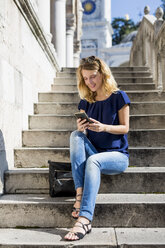 Italien, Udine, lächelnde blonde Frau sitzt auf einer Treppe und schaut auf ihr Handy - MAUF000839