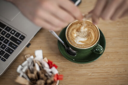 Hand gießt Zucker in eine Tasse Cappuccino neben dem Laptop - ZEF009853