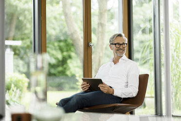 Smiling man with tablet sitting on chair in his living room - SBOF000182