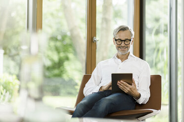 Smiling man sitting on chair in his living room using tablet - SBOF000183