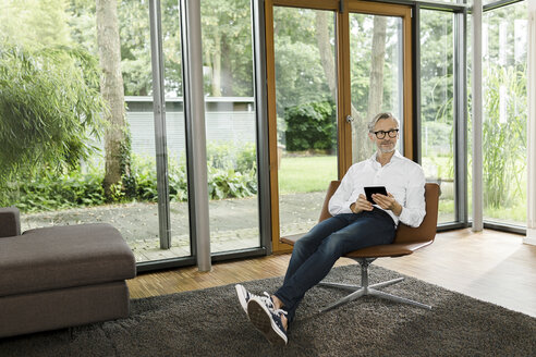 Mann sitzt mit Tablet auf einem Stuhl in seinem Wohnzimmer - SBOF000185