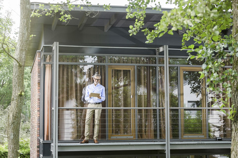 Mann steht auf dem Balkon seines Hauses und benutzt ein Tablet, lizenzfreies Stockfoto
