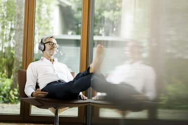 Man sitting on chair in his living room listening music with headphones - SBOF000198