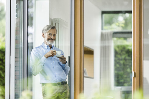 Lächelnder Mann mit einer Tasse Kaffee, der in seinem Haus steht und durch das Fenster schaut - SBOF000230