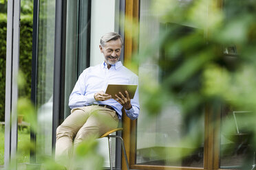 Mann sitzt auf einem Stuhl vor seinem Haus und benutzt ein Tablet - SBOF000231