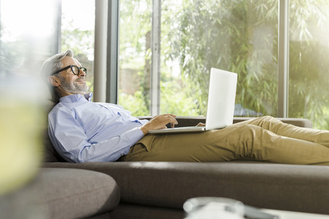 Lächelnder Mann, der in seinem Wohnzimmer auf der Couch liegt und einen Laptop benutzt, lizenzfreies Stockfoto