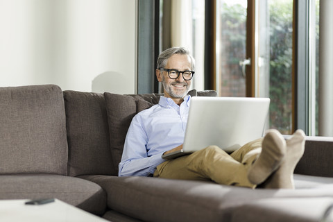 Smiling man sitting on couch in his living room using laptop stock photo