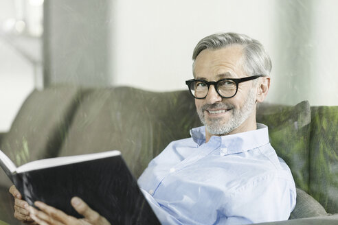 Porträt eines lächelnden Mannes, der mit einem Buch auf der Couch sitzt - SBOF000243