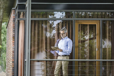 Mann steht auf dem Balkon seines Hauses und schaut auf ein Tablet - SBOF000248