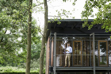 Man standing on balcony of his house looking at tablet - SBOF000249