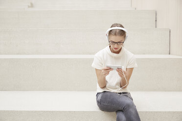 Junge Frau mit Kopfhörern sitzt auf einer Treppe und schaut auf ihr Smartphone - FMKF003090