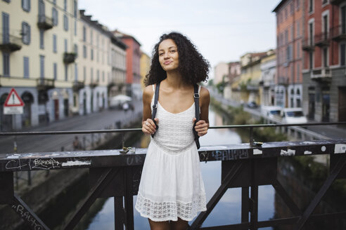 Italy, Milan, portrait of smiling young woman with backpack wearing white summer dress standing on a bridge - MRAF000124