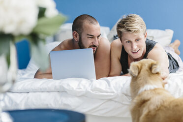 Schwules Paar mit Hund im Bett liegend, mit Laptop - MADF001144