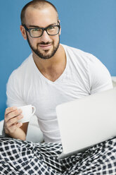 Gay man sitting on bed, using laptop and drinking coffee - MADF001147