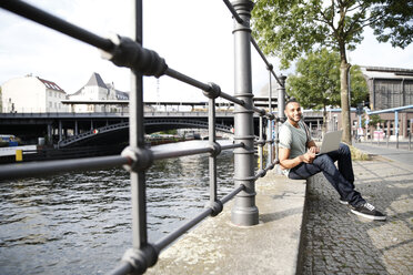 Junger Mann sitzt am Fluss und benutzt einen Laptop - FKF002041