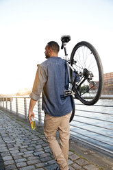 Young man walking by river, carrying bicycle - FKF002054