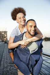 Young man carrying woman piggyback by river - FKF002074