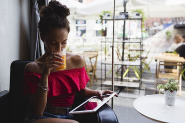 Junge Frau trinkt ein Glas Orangensaft in einem Café und schaut auf ein Tablet - MRAF000147