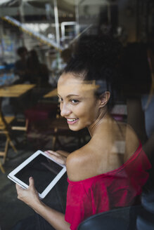 Lächelnde junge Frau mit Tablet, die durch das Fenster eines Cafés schaut - MRAF000150