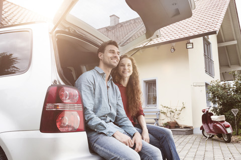 Lächelndes Paar sitzt im Kofferraum eines Autos vor einem Haus, lizenzfreies Stockfoto