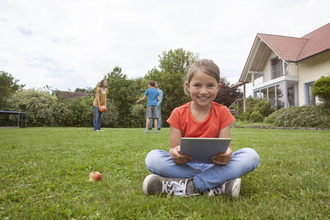 Lächelndes Mädchen sitzt im Garten und benutzt ein Tablet mit seiner Familie im Hintergrund, lizenzfreies Stockfoto