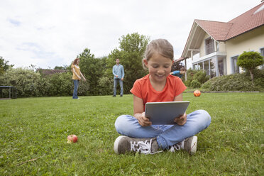 Smiling girl sitting in garden using tablet with family in background - RBF005139
