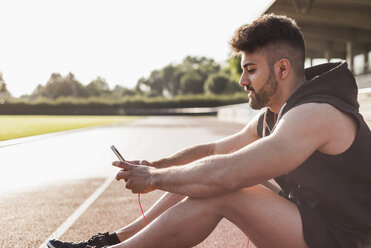 Young man with cell phone sitting on tartan track - UUF008280