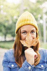 Porträt einer lächelnden jungen Frau mit gelber Kappe, die den Mund mit einem Herbstblatt bedeckt - EBSF001674