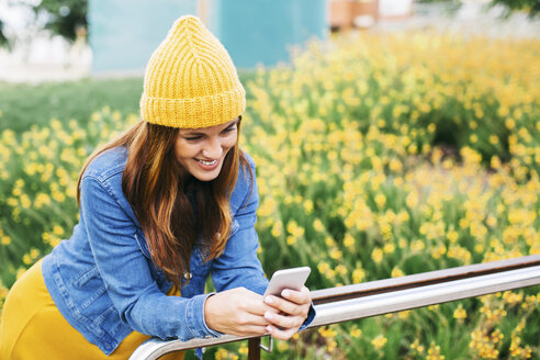 Lächelnde junge Frau mit gelber Mütze, die auf ihr Mobiltelefon schaut - EBSF001677